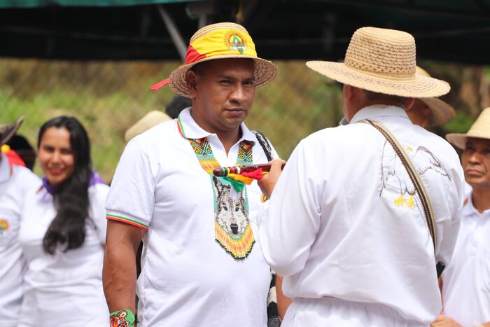A man in a white shirt, hat and ceremonial necklace is receiving an item from another person
