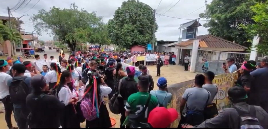 a group of around 50 people stand with banners around a central person speaker. they are all next to a small building with a big gate next to it